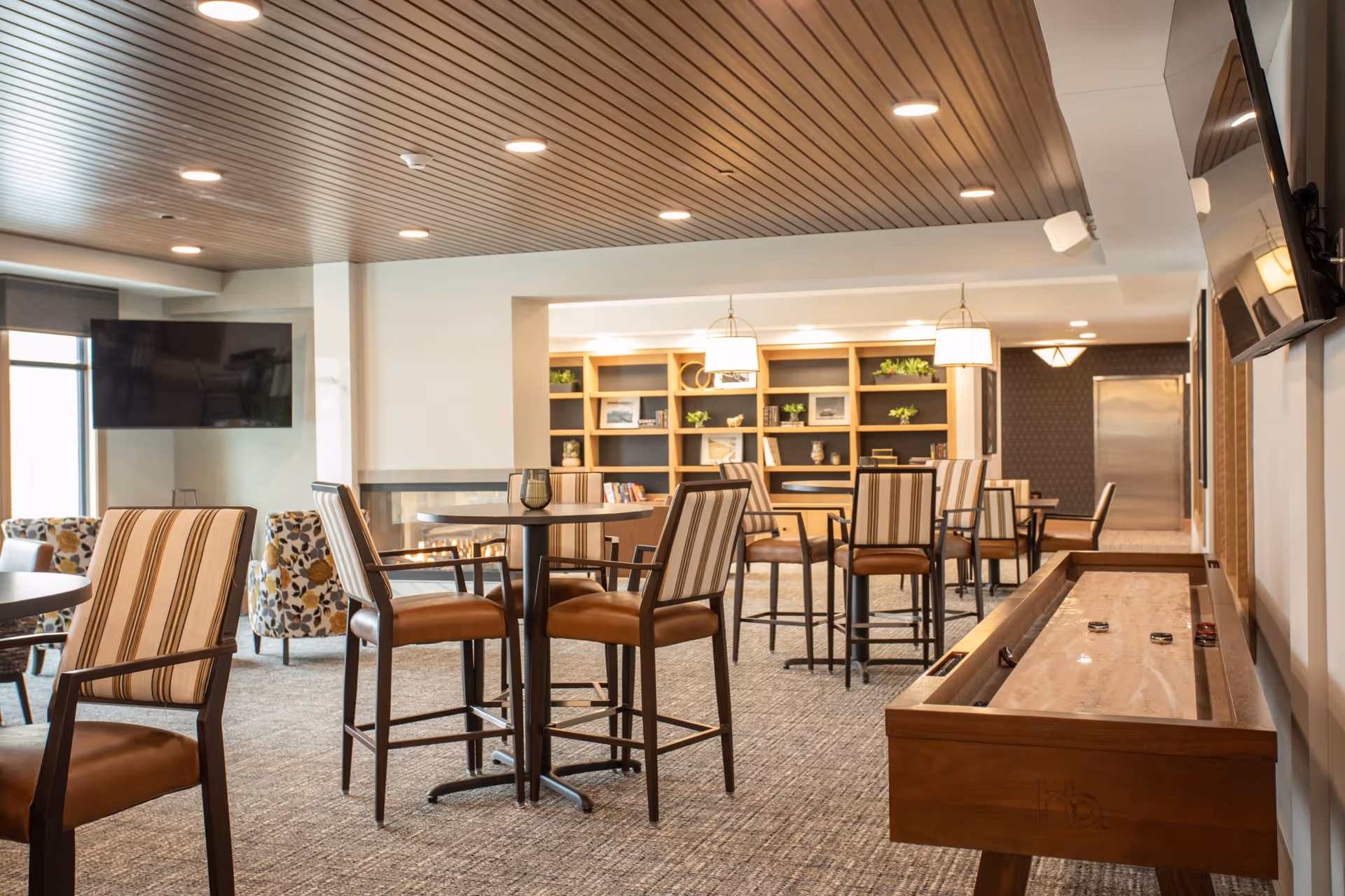 A spacious and well-lit common area with high tables and chairs, a shuffleboard table, a fireplace, a large wall-mounted TV, and a bookshelf with decorative items and plants in the background.
