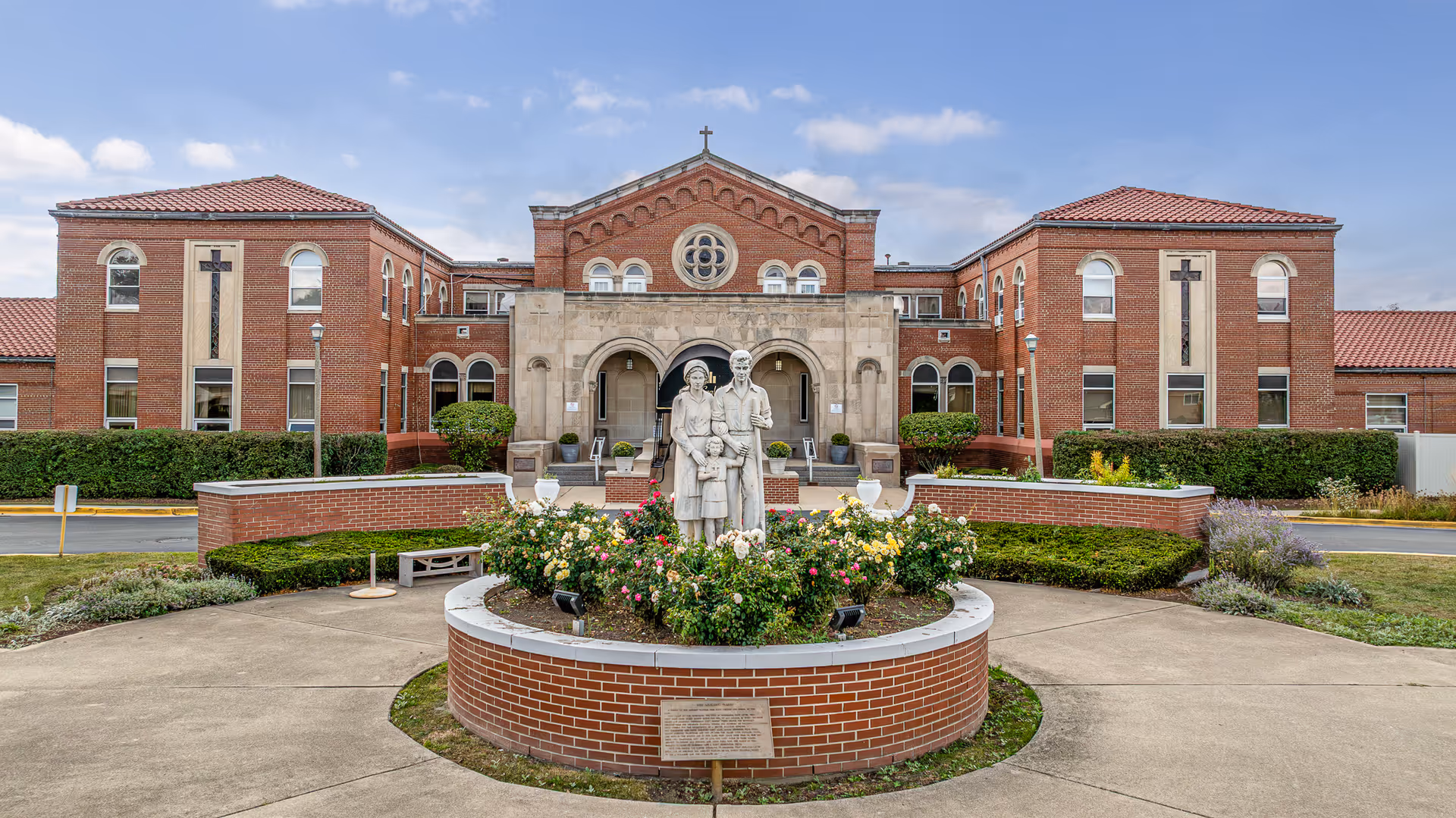 Brick senior living facility with circular flower bed, statue, and front entrance.