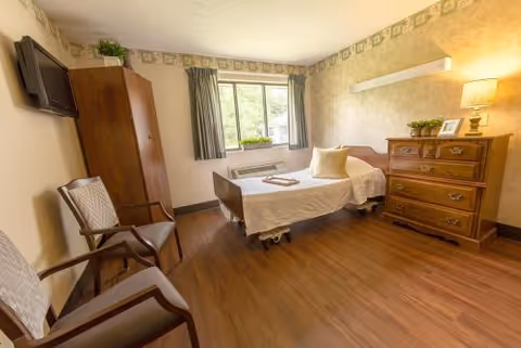 A well-lit room with a single hospital-style bed, two chairs, a wooden dresser with a lamp and framed photo, a wooden wardrobe, and a window with green curtains overlooking greenery outside. The room has wooden flooring and light-colored walls with a decorative border near the ceiling.