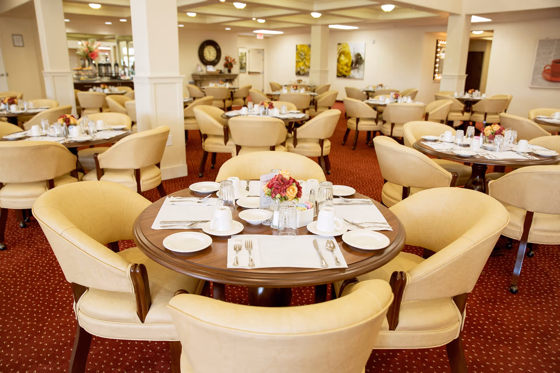 A bright and spacious dining room with multiple round wooden tables set for meals. Each table is surrounded by beige upholstered chairs and has white plates, cups, silverware, and small floral centerpieces. The room has a red carpet with a subtle pattern, cream-colored walls, and decorative artwork. There is a clock and a buffet area visible in the background.