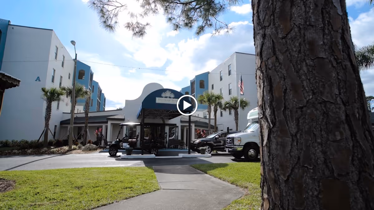 Exterior view of Fellowship at The Fairway Assisted Living Community building with a driveway, golf carts, palm trees, and a large tree trunk in the foreground under a partly cloudy sky.