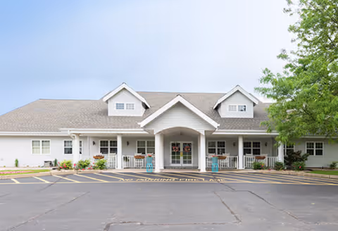 Front entrance of a single-story white senior living building with a covered porch and parking area.