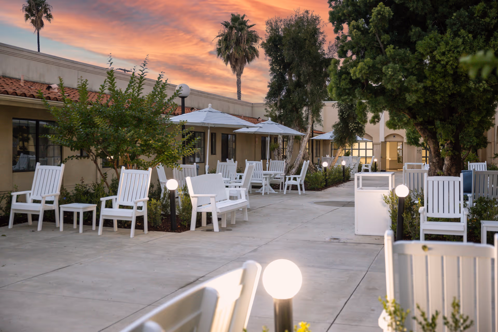 Outdoor courtyard with white chairs, tables, umbrellas, and trees at a senior living center.