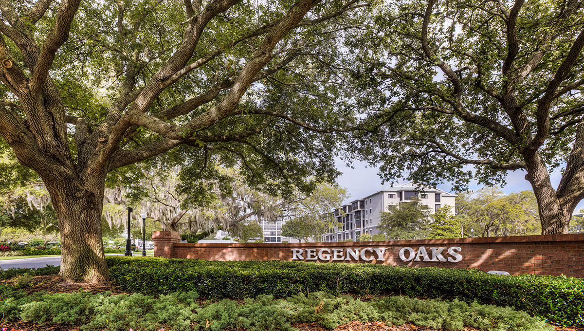 Entrance sign for Regency Oaks Health Center made of brick with white letters, surrounded by large oak trees and greenery, with a multi-story building visible in the background under a partly cloudy sky.