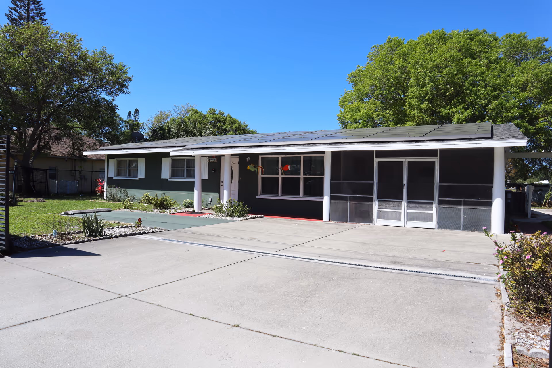 Single-story green and white house with a screened porch, solar panels on the roof, and a large concrete driveway under a clear blue sky.