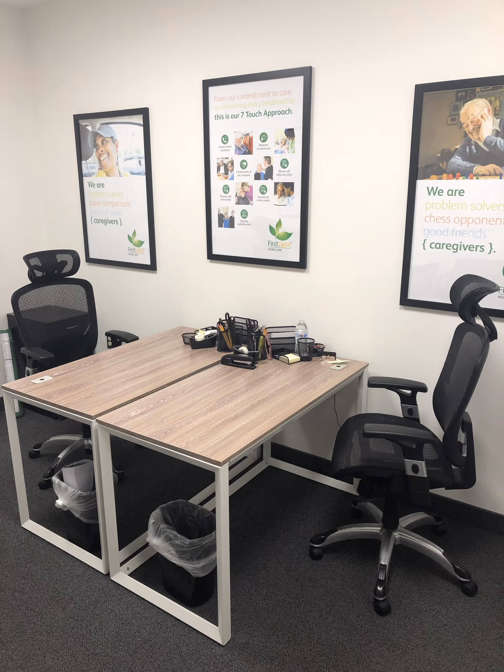 Office workspace with two desks, chairs, and framed FirstLight posters on the wall.