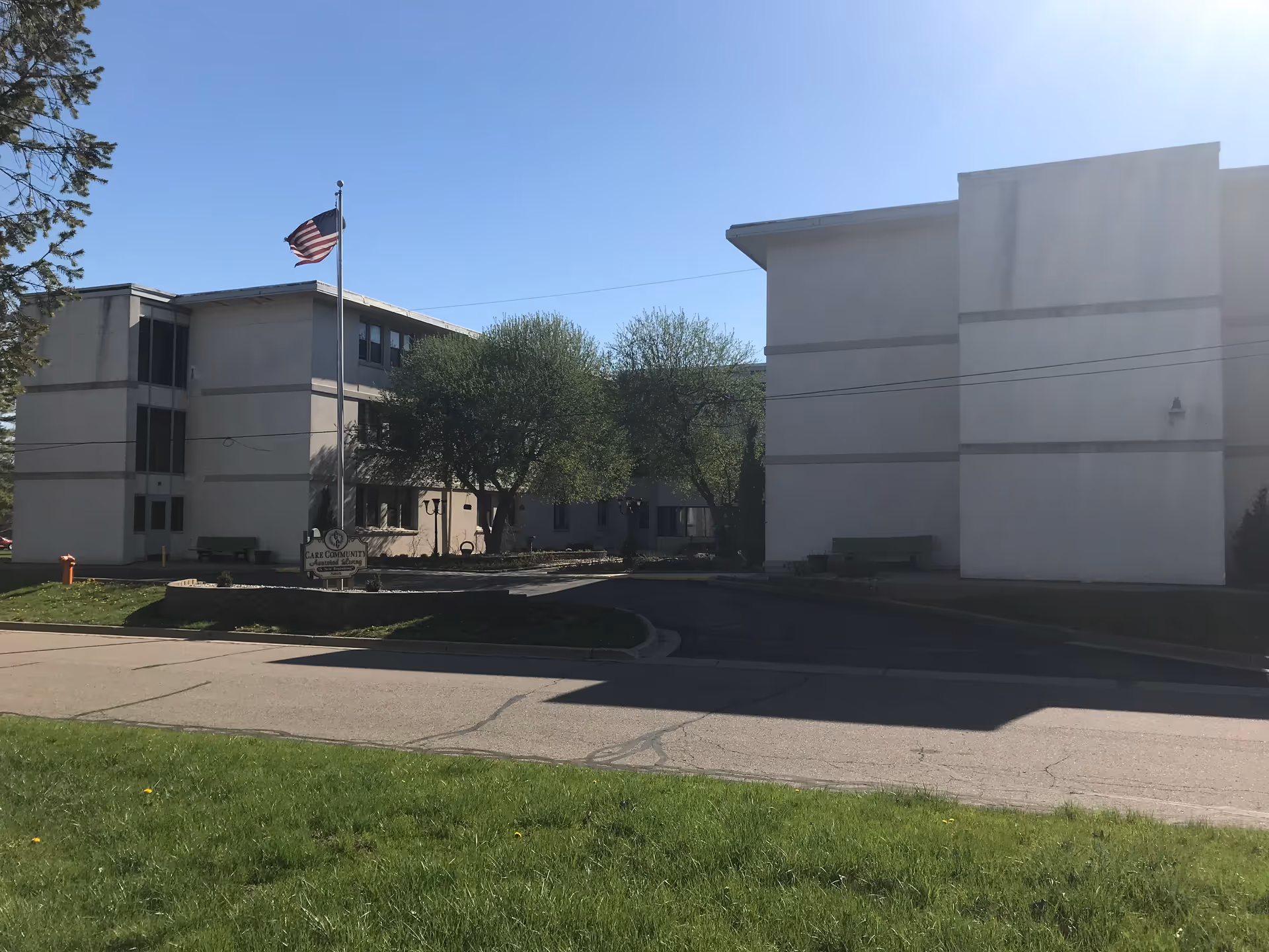 Exterior view of a multi-story senior living facility building with an American flag on a flagpole in front. There are trees and a small garden area between the building sections, and a sign that reads 'Care Community Assisted Living'. The sky is clear and blue.