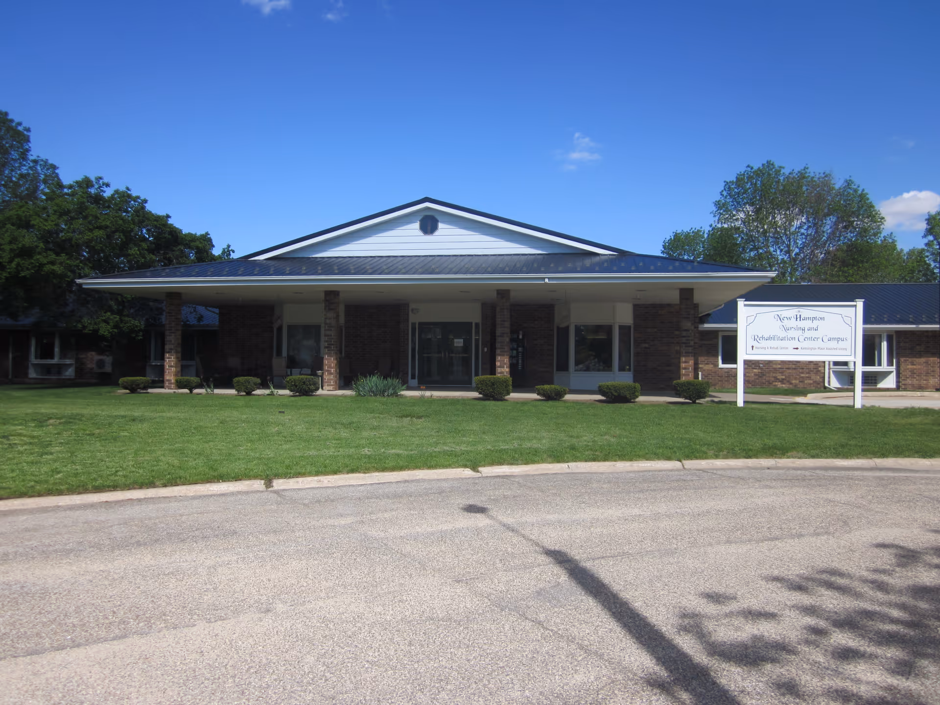 Front exterior of a single-story brick nursing and rehabilitation building with a covered entrance, lawn, and a sign near the driveway.