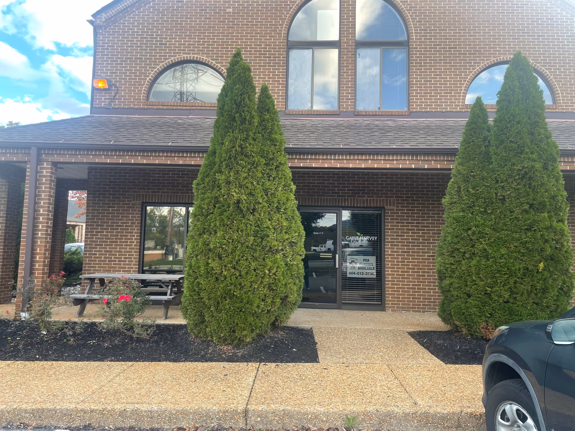 Brick building front with arched windows, two tall evergreens, and a glass entrance door.