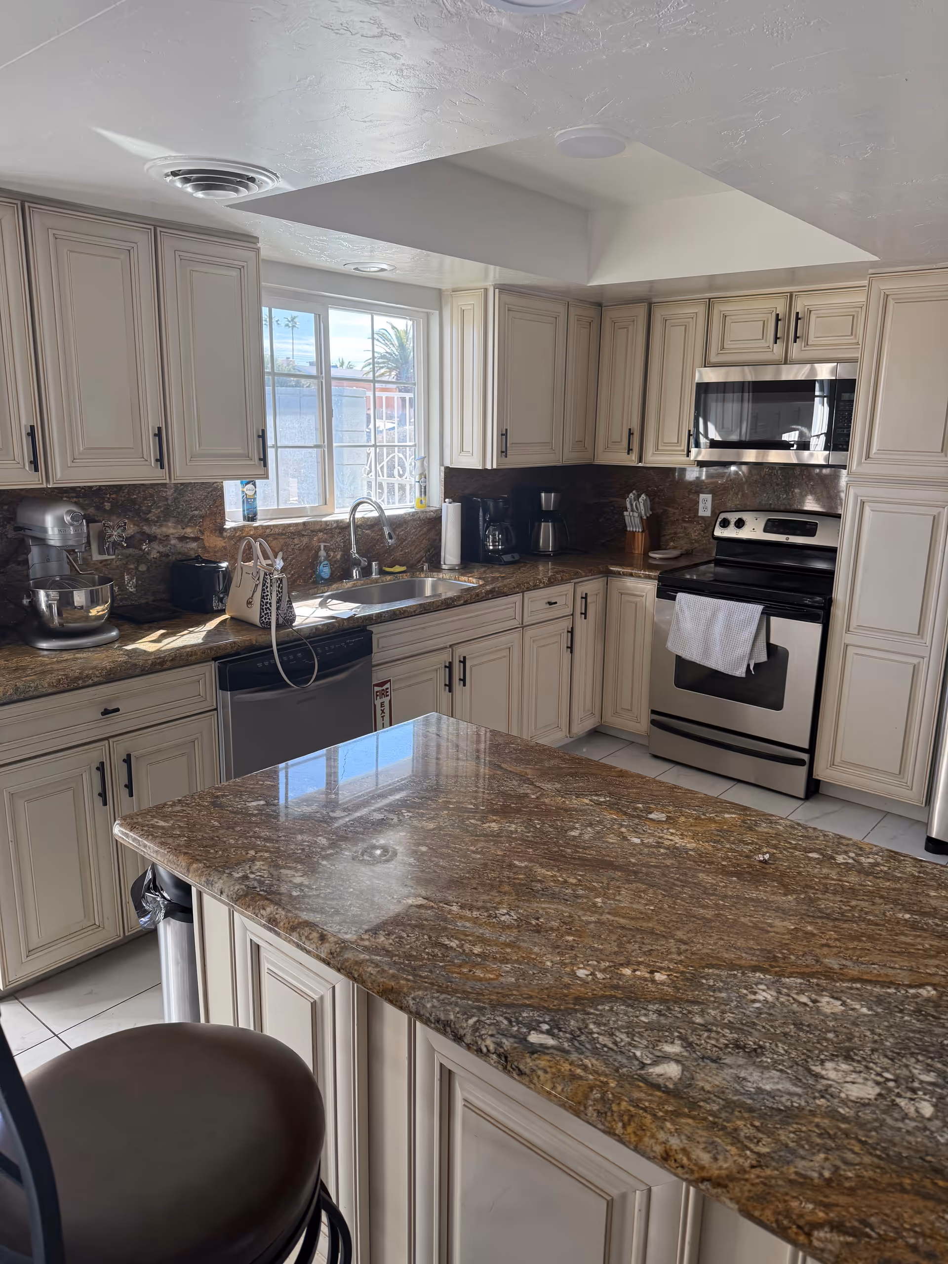 Bright kitchen with beige cabinets, granite countertops, island, and stainless steel stove and appliances by window.