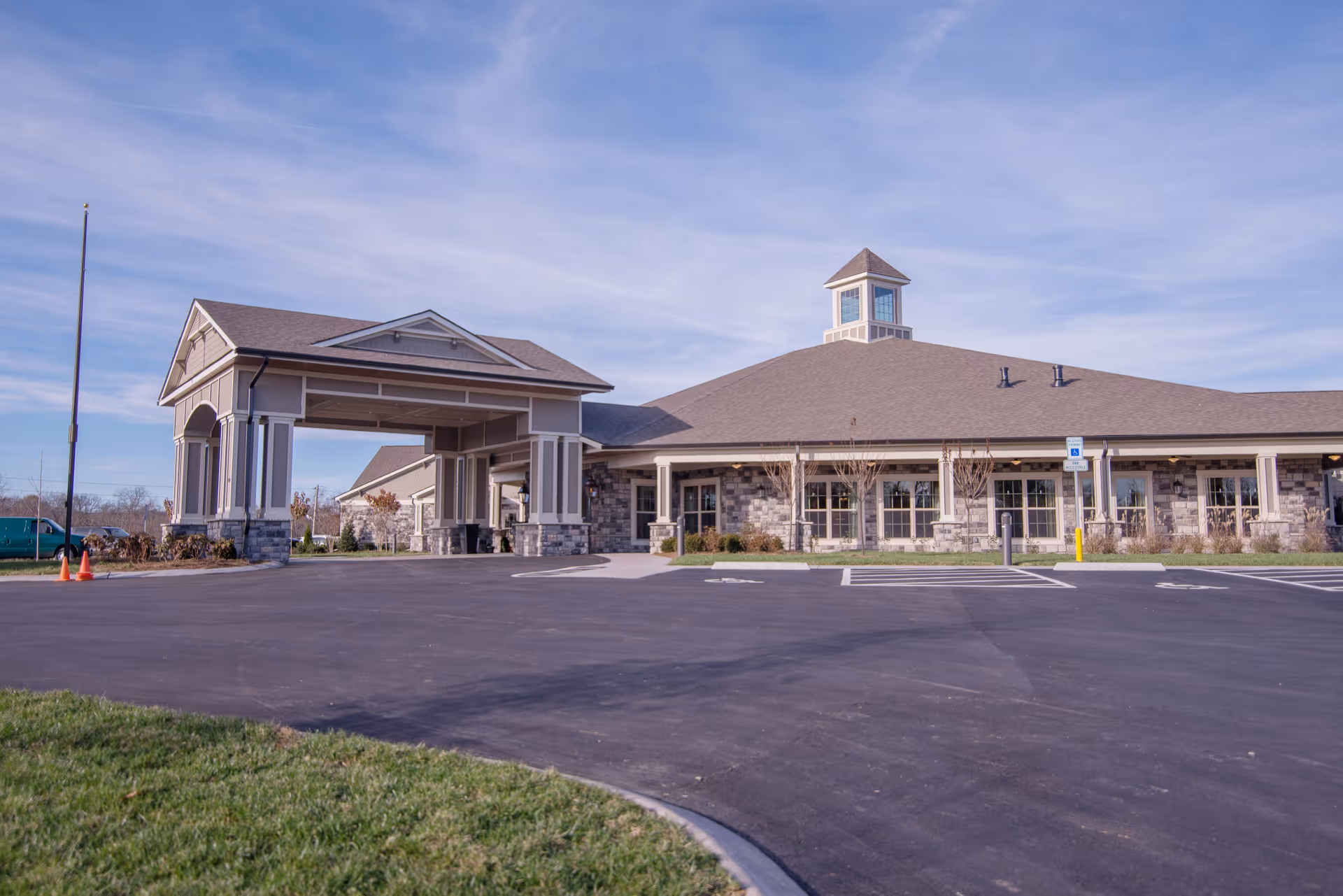 Exterior view of a single-story senior living facility building with a covered entrance and a cupola on the roof, surrounded by a paved parking lot and some grass under a partly cloudy sky.