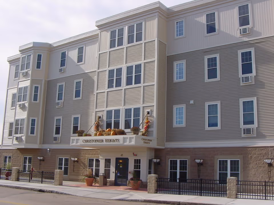 Exterior front view of a multi-story senior living facility named Christopher Heights. The building has beige and light brown siding with multiple windows and air conditioning units. The entrance is decorated with autumn-themed items including pumpkins and dried corn stalks.