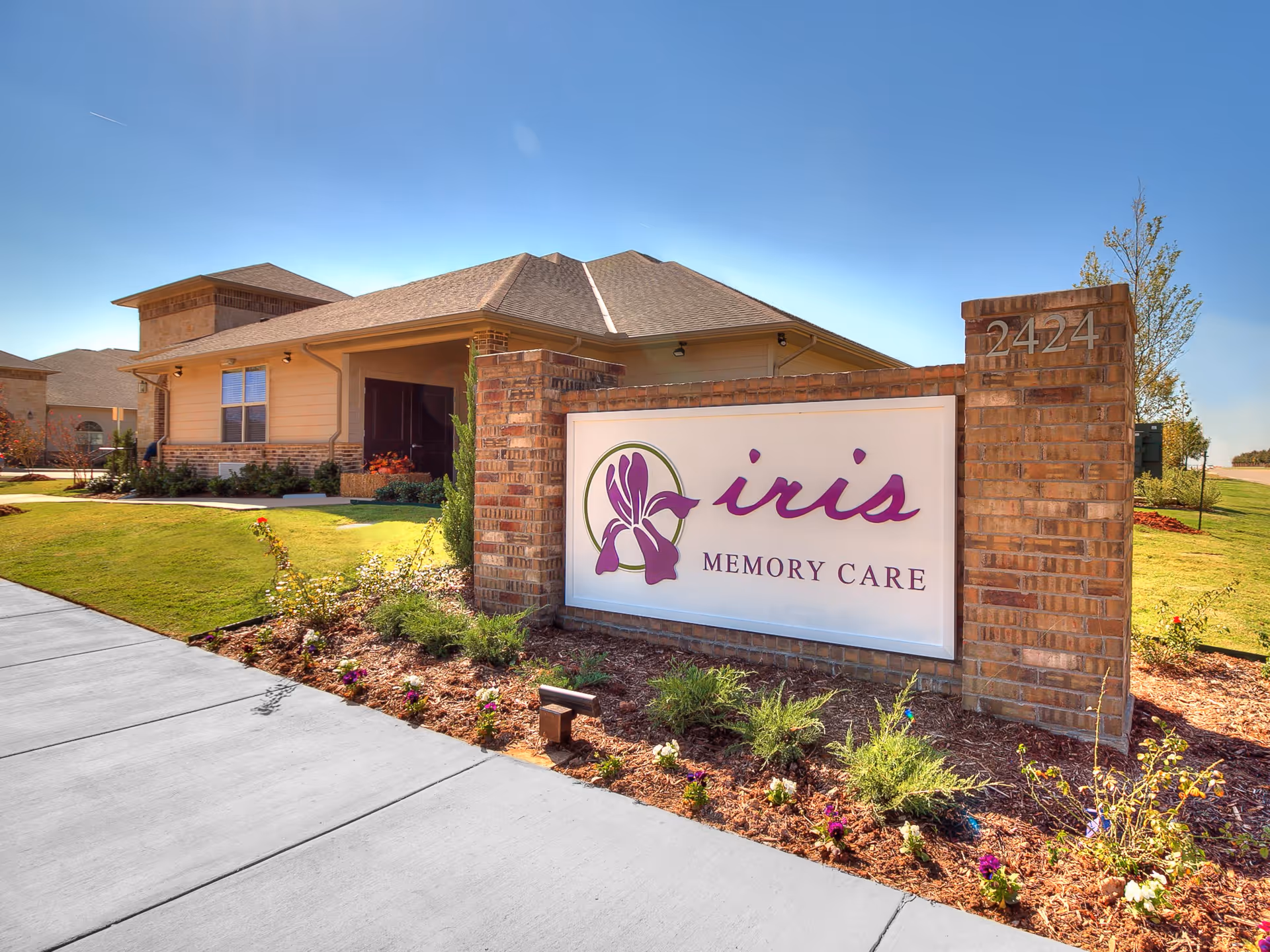 Exterior view of Iris Memory Care of Edmond building with a brick sign displaying the facility name and address number 2424, surrounded by landscaped grass and flowers under a clear blue sky.