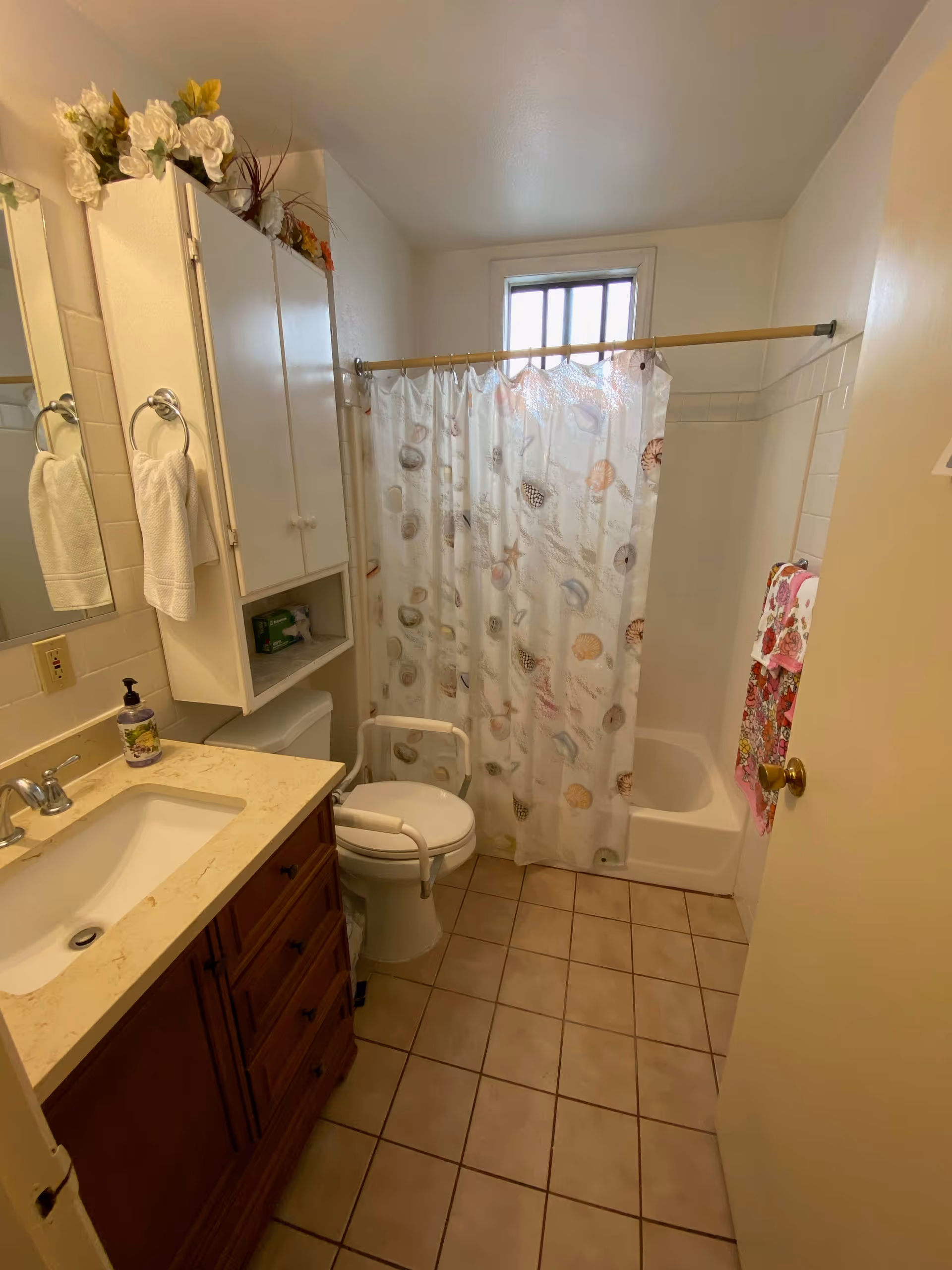 Small tiled bathroom with a sink vanity, cabinet, toilet with safety rails, and a bathtub with a patterned shower curtain.