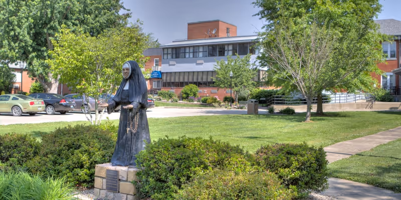 Statue of a nun holding a rosary on the lawn in front of a multi-story healthcare building with trees and parked cars.