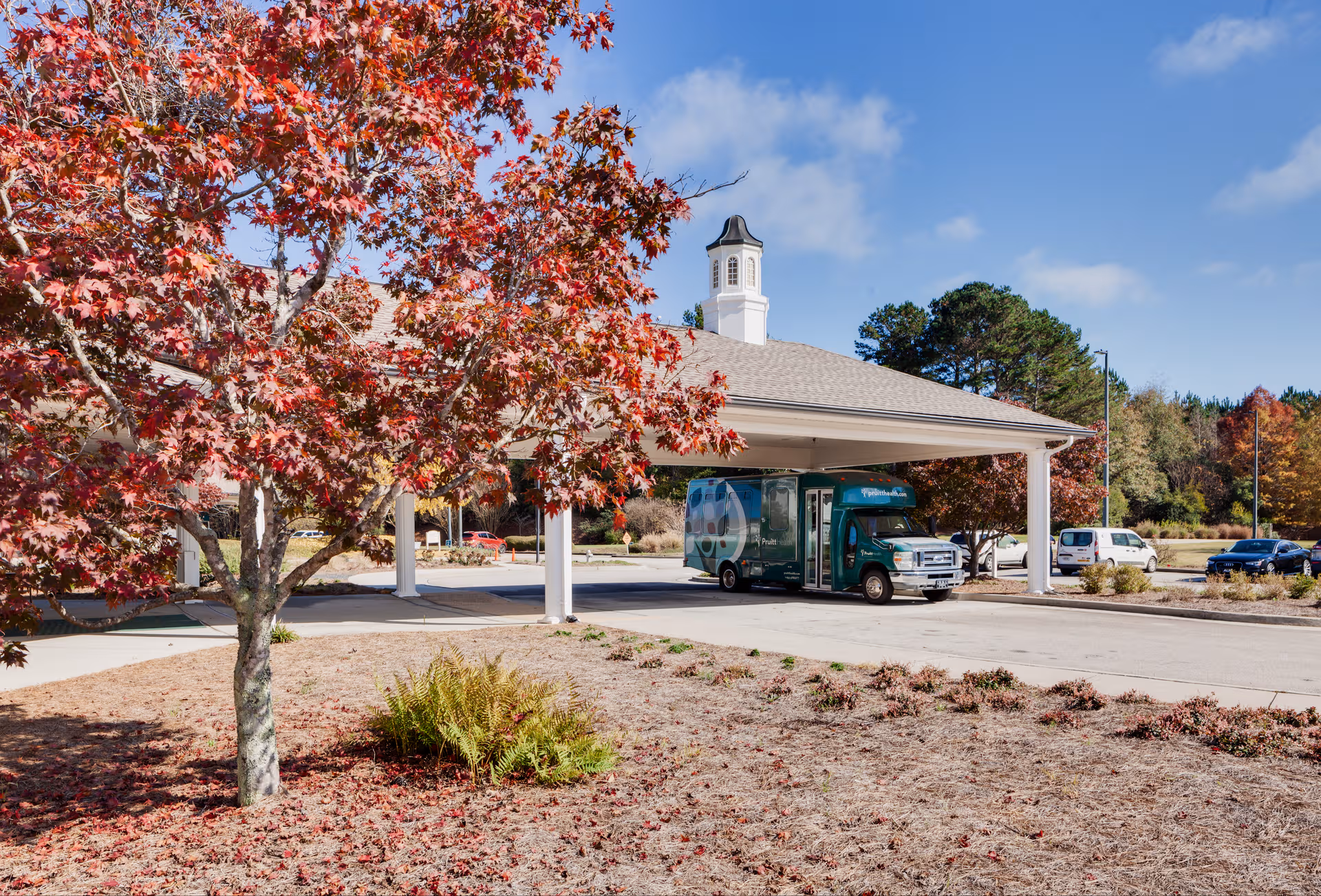 Red-leaved tree foreground frames a building entrance with a covered driveway and truck.