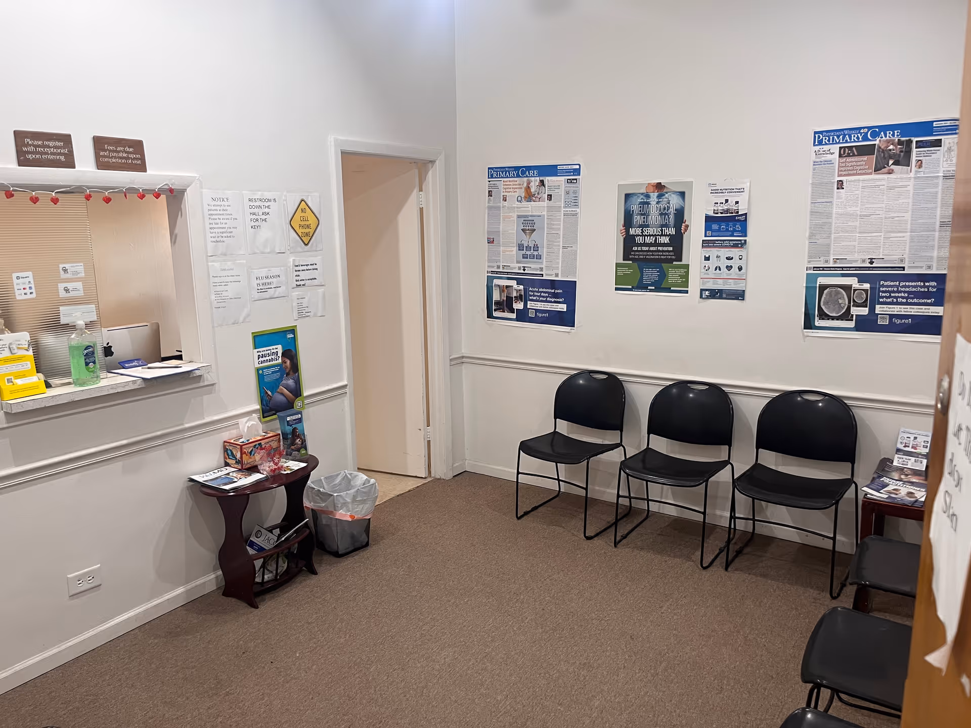 A waiting area with beige carpet, three black chairs, posters on walls, and a reception window.