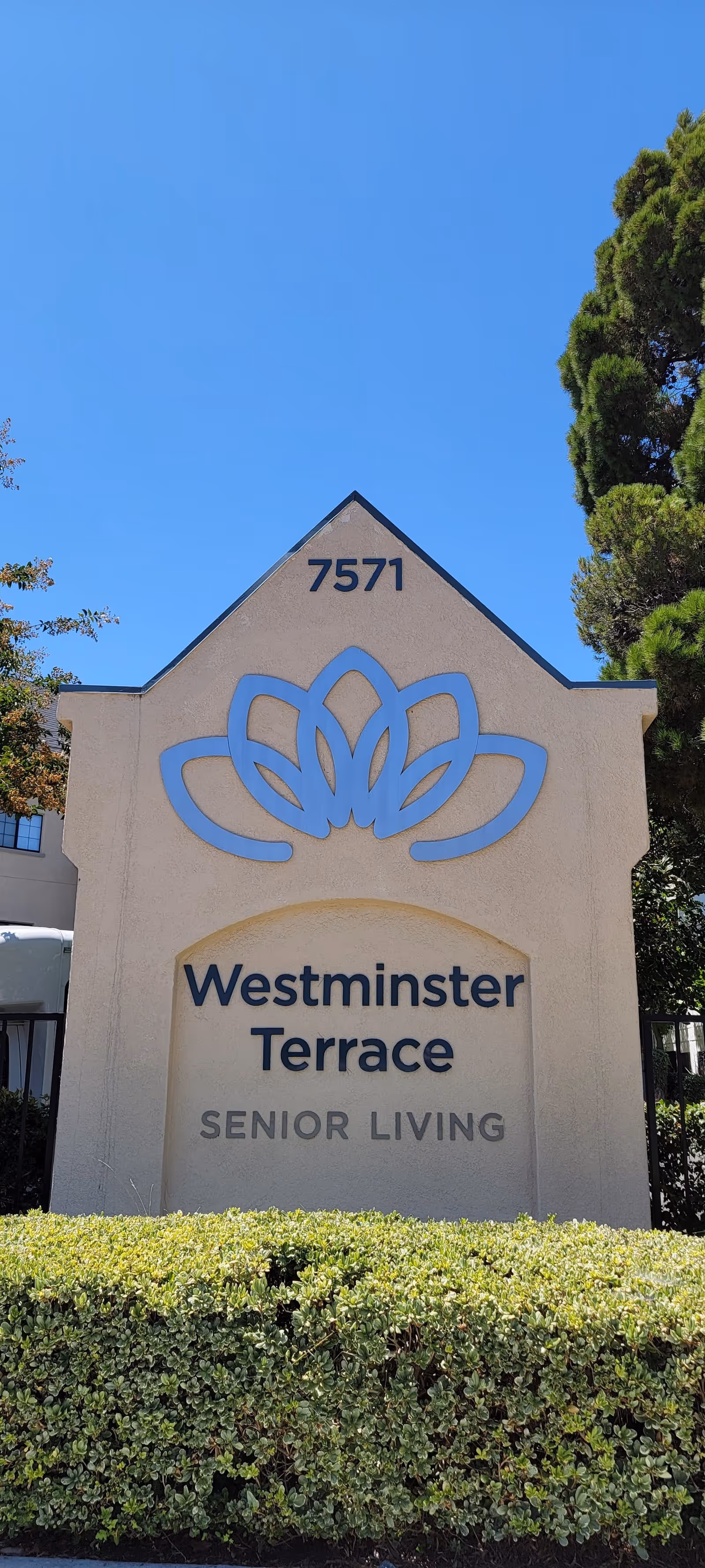 Outdoor sign for Westminster Terrace Senior Living with the address number 7571 displayed above a blue floral logo, surrounded by greenery and trees under a clear blue sky.