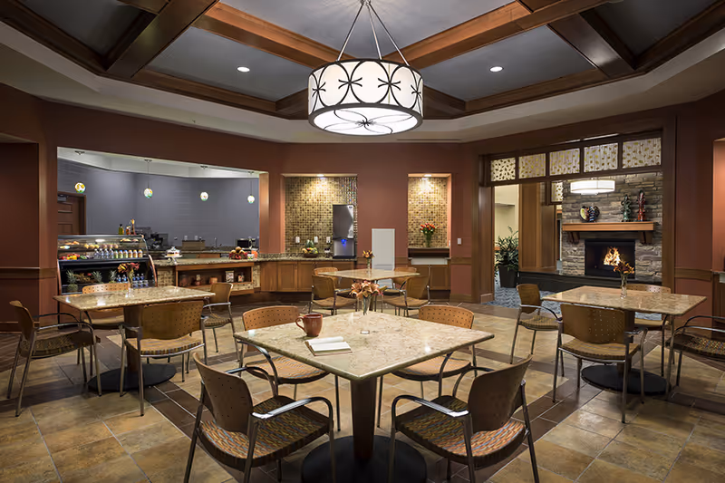 A warm, well-lit dining area with marble-top tables and chairs, a serving counter and a fireplace in the background.