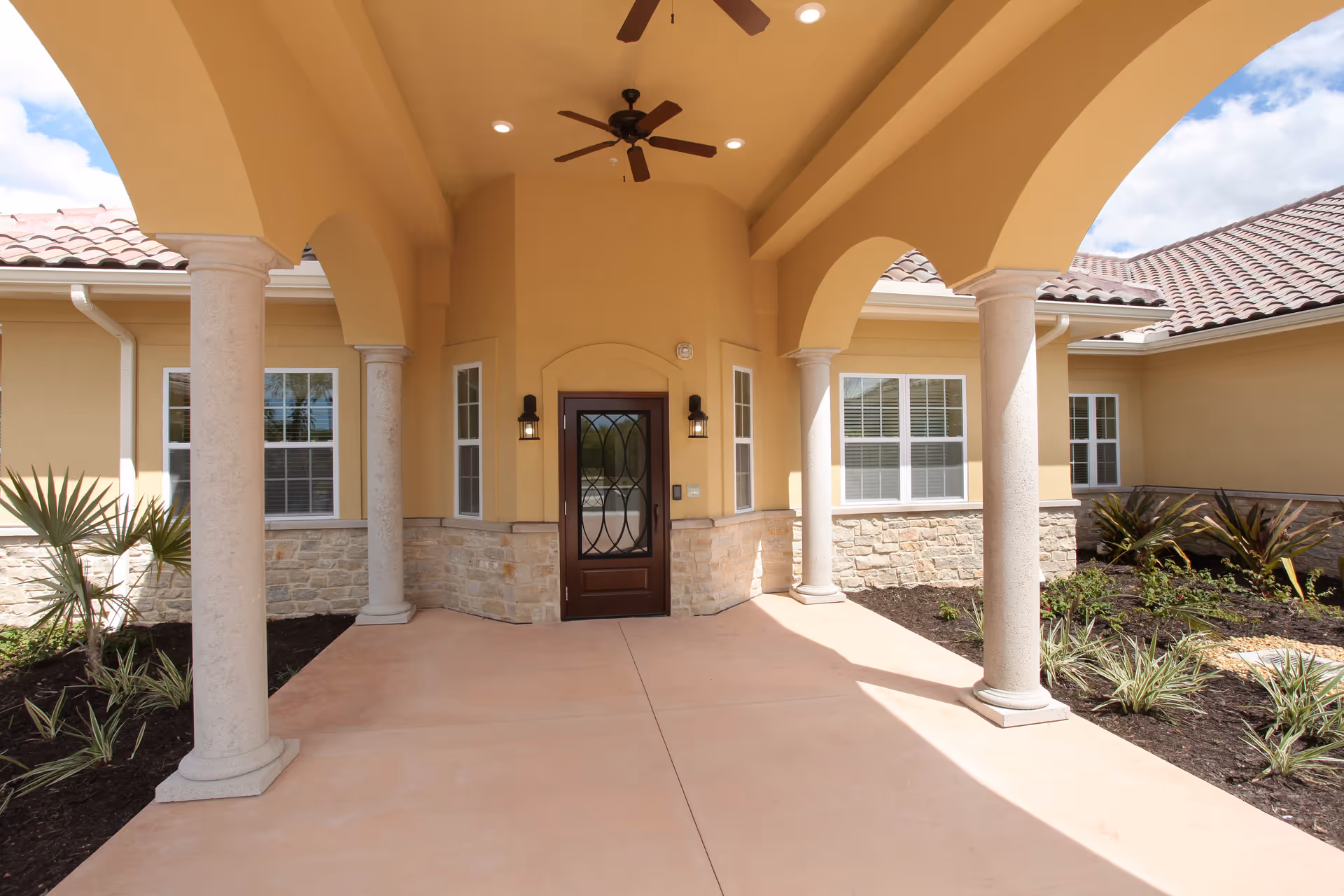Covered entrance area of a building with beige walls, stone accents, and a dark wooden door with decorative glass. The entrance is framed by four stone columns supporting arches and a ceiling with two ceiling fans and recessed lighting. There are windows on either side of the door and landscaped garden beds with plants along the sides.