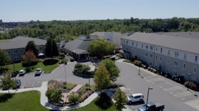 Aerial view of Benedictine Living Community-St. Joseph showing multiple connected buildings surrounded by trees and greenery. There is a circular driveway with parked cars and landscaped areas with bushes and trees. The sky is clear and the setting appears peaceful.