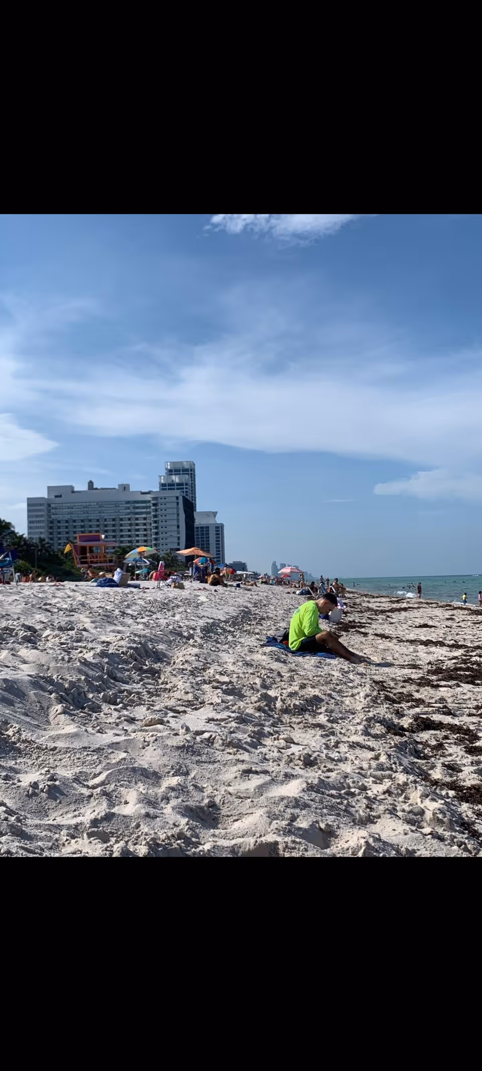 Sunlit beach scene with people on sand, umbrellas, and distant city skyline.