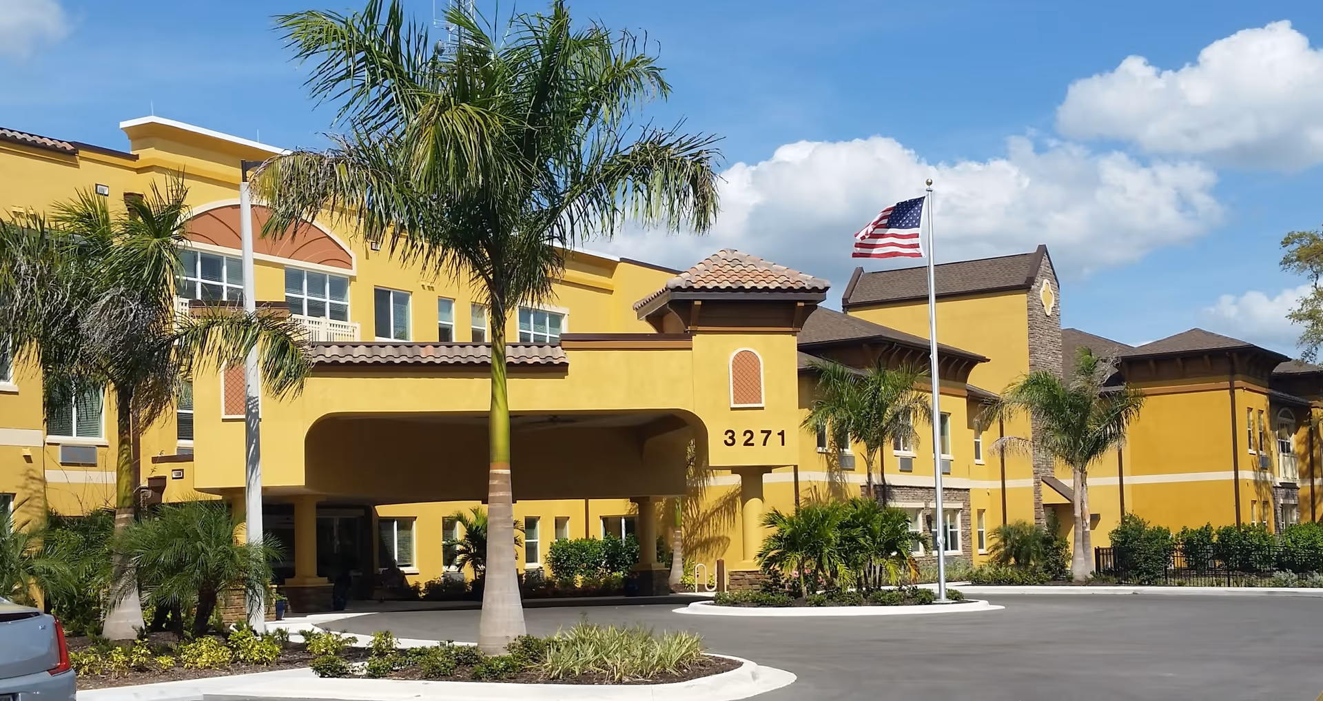 Exterior view of a yellow senior living facility building with palm trees and an American flag in front. The building has a covered entrance with the number 3271 displayed on it, under a clear blue sky with some clouds.