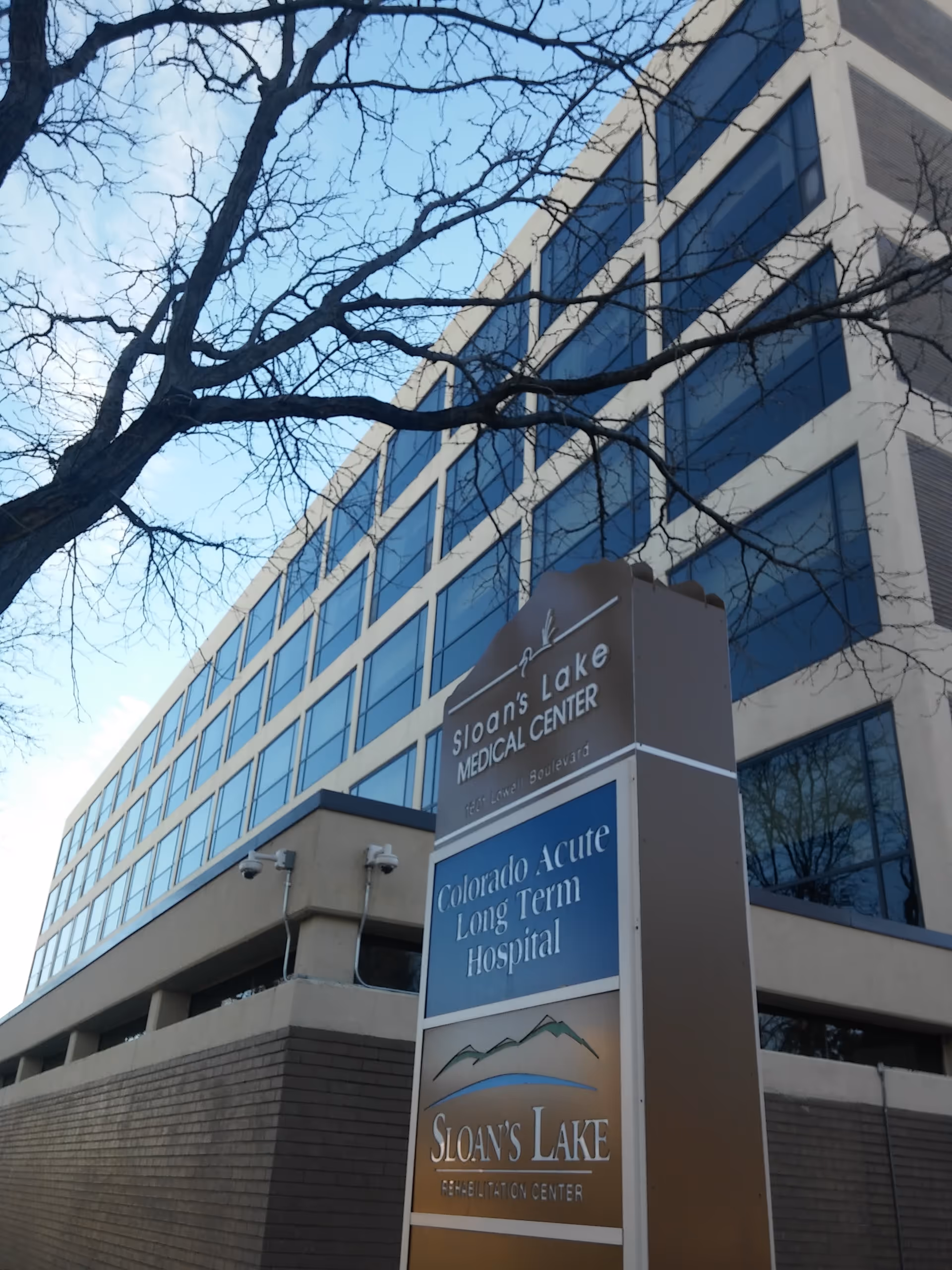 Tall glass-front medical center building with Sloan's Lake sign and leafless trees in foreground.