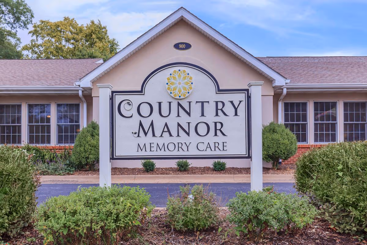Exterior view of a building with a large sign in front that reads 'Country Manor Memory Care'. The building has multiple windows and is surrounded by bushes and greenery under a partly cloudy sky.