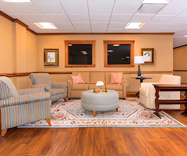 A cozy living room area with a beige sofa adorned with pink pillows, two armchairs with striped and solid upholstery, a round ottoman in the center on a patterned rug, wooden flooring, two framed pictures on the wall, and a table lamp on a side table.