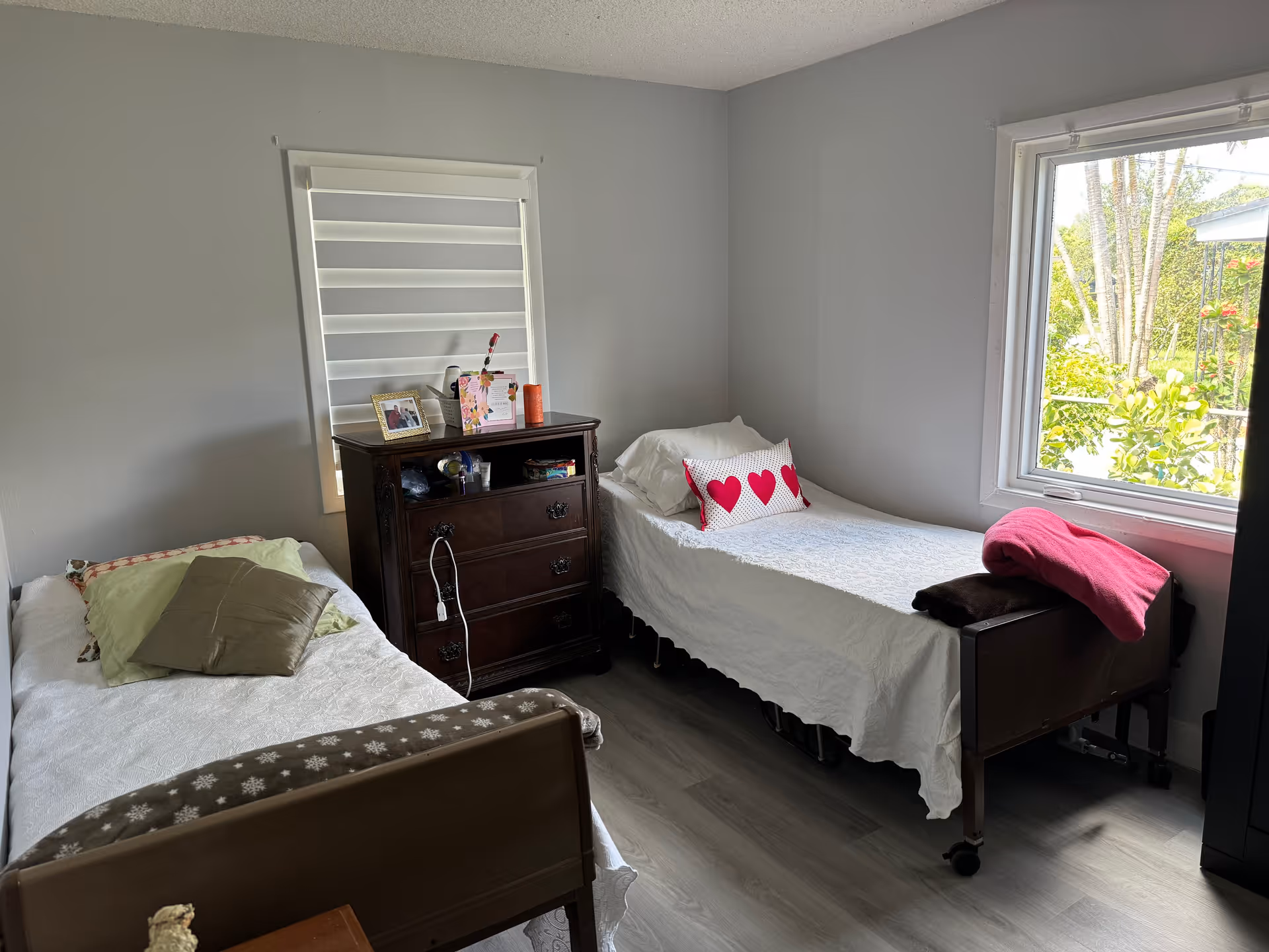 A small bedroom with two single beds, each covered with white bedspreads. One bed has a green pillow and a brown pillow, while the other has a white pillow and a decorative pillow with three red hearts. Between the beds is a dark wooden dresser with framed photos, a flower, and other small items on top. A large window on the right side lets in natural light and shows greenery outside. The walls are painted light gray and the floor is a light wood laminate.