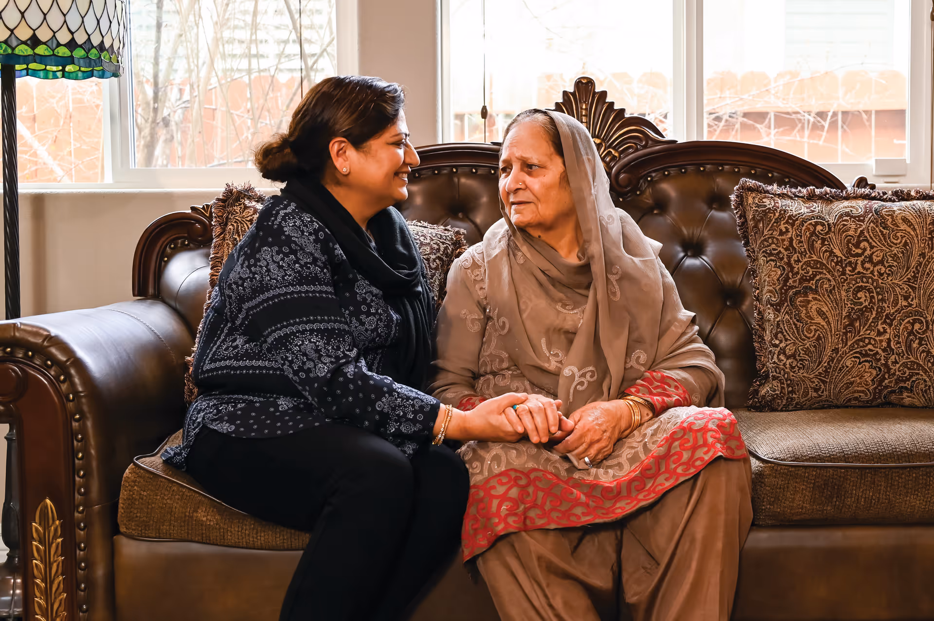 Two women hold hands and chat on a leather couch in a cozy living room.
