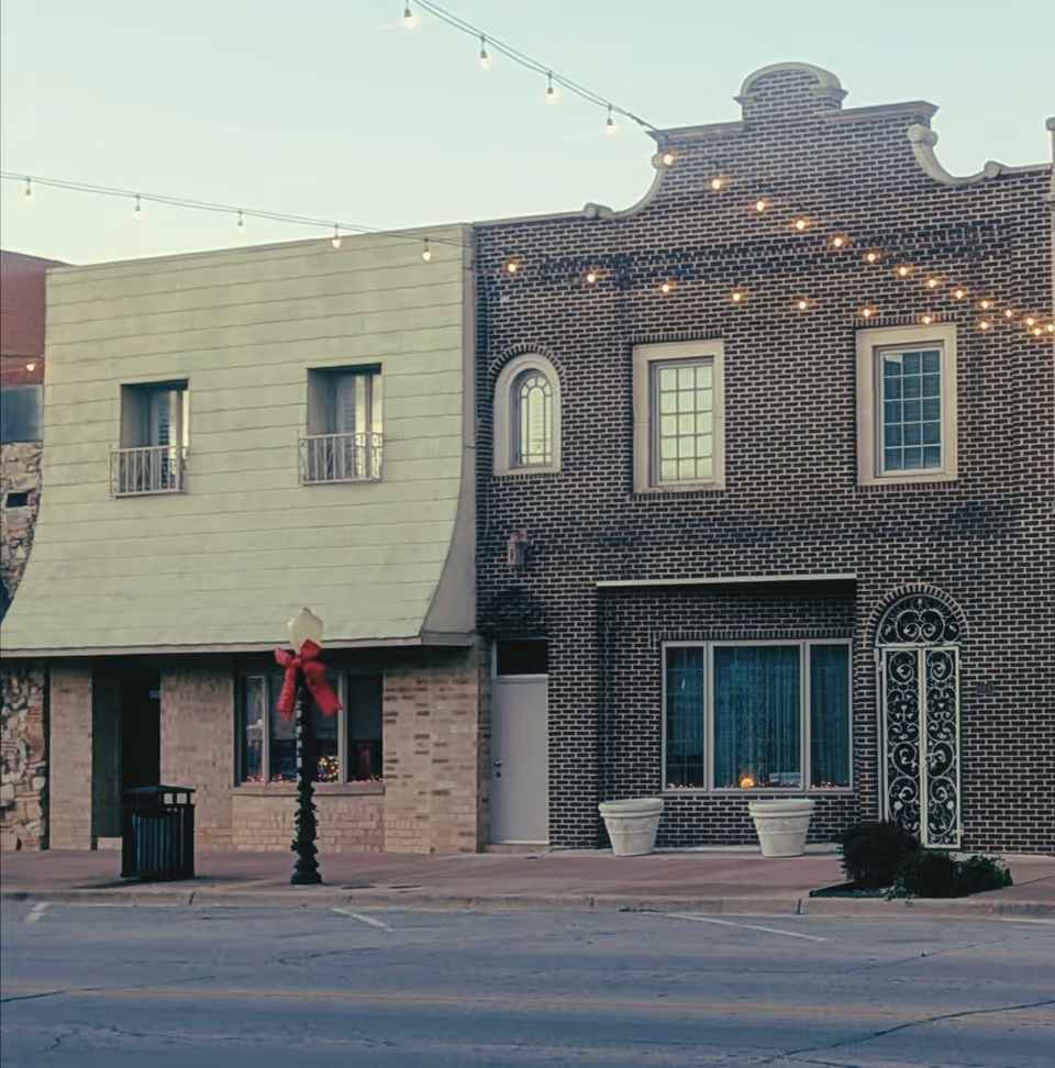 Exterior view of two connected brick buildings on a street with string lights overhead.