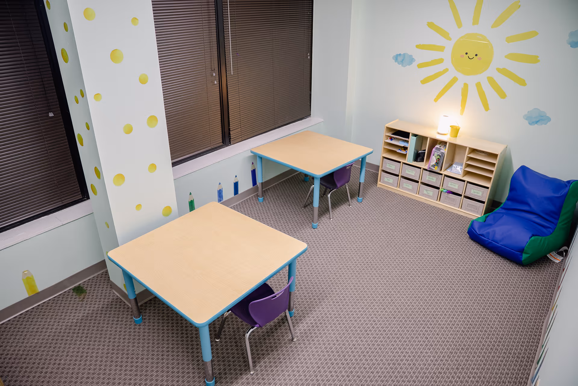 Bright children's activity room with two small tables, colorful chairs, cubby storage, and sun mural.