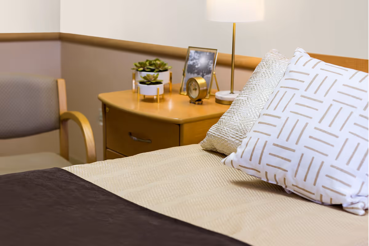 Close-up view of a neatly made bed with patterned pillows and a beige bedspread. Next to the bed is a wooden nightstand holding a small potted plant, a framed photo, a round clock, and a lamp. A cushioned chair with wooden armrests is positioned beside the nightstand against a beige and light brown wall.