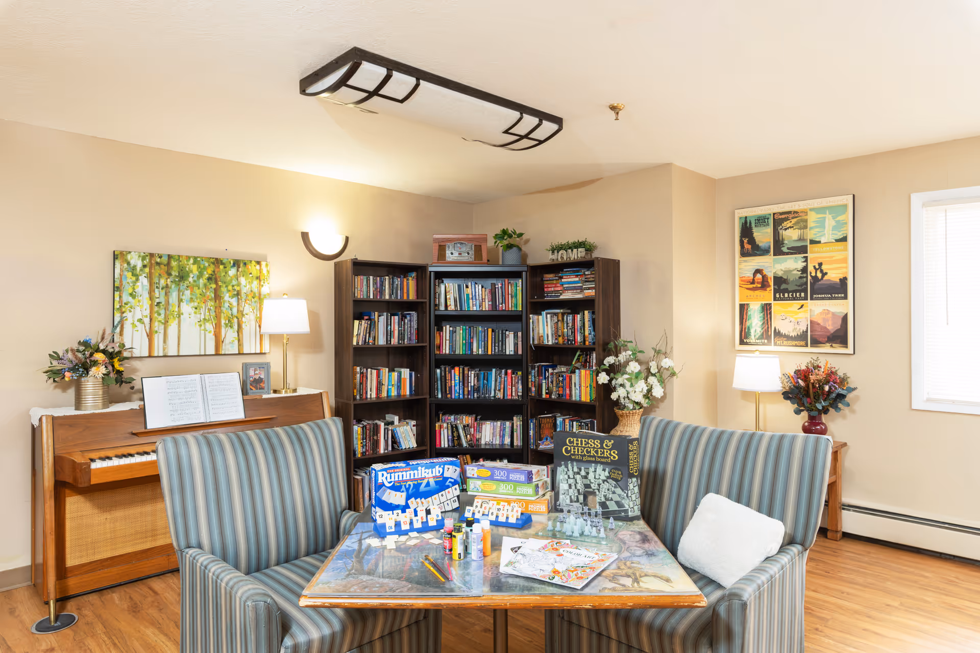 Cozy communal living room with a piano, bookshelves, game table, and striped chairs.