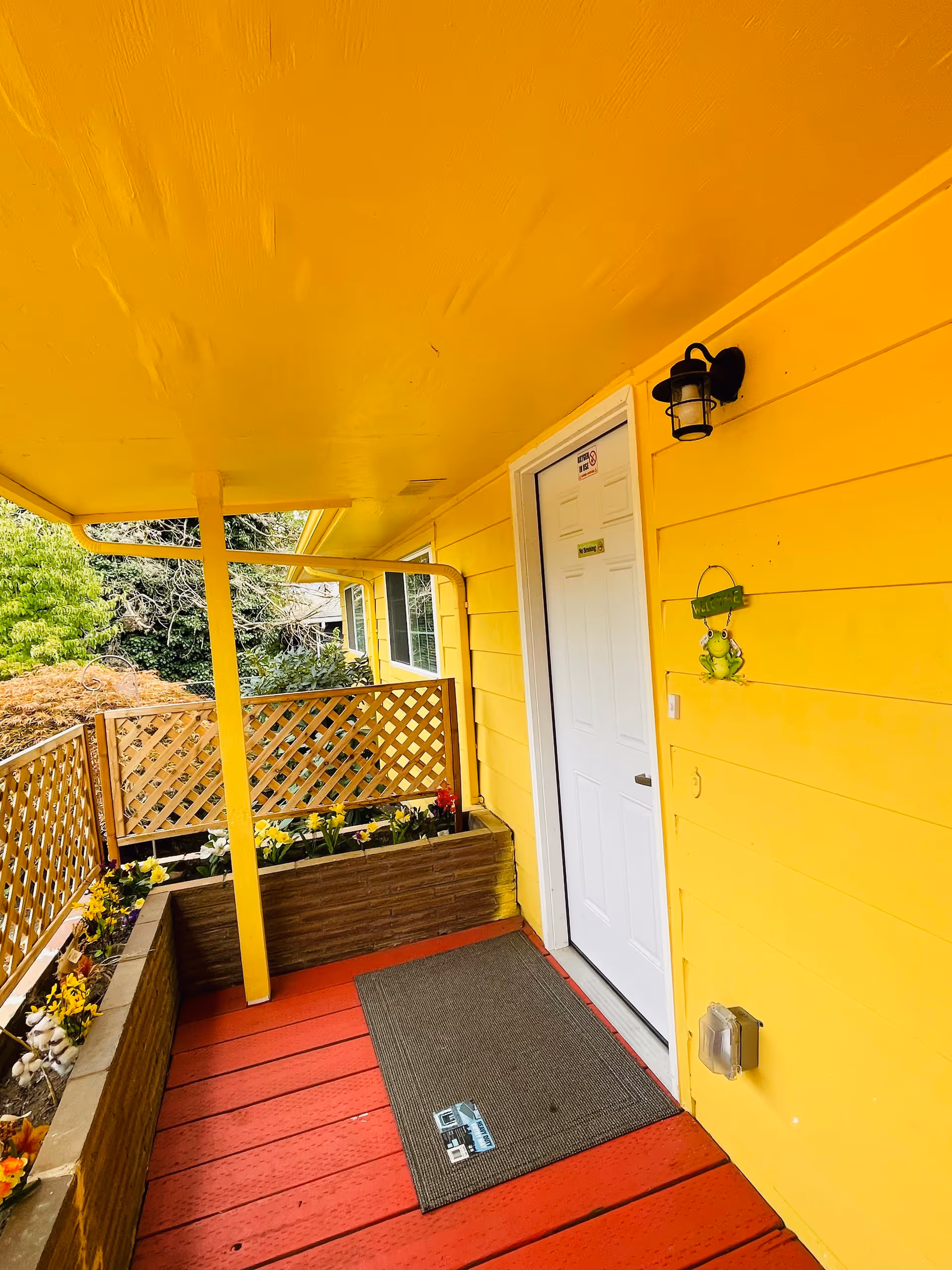 Covered porch area with bright yellow walls and ceiling, a white door with a small sign, a black outdoor wall light, a welcome sign with a frog decoration, red wooden floorboards, a gray doormat, and flower beds with various plants along the edges.