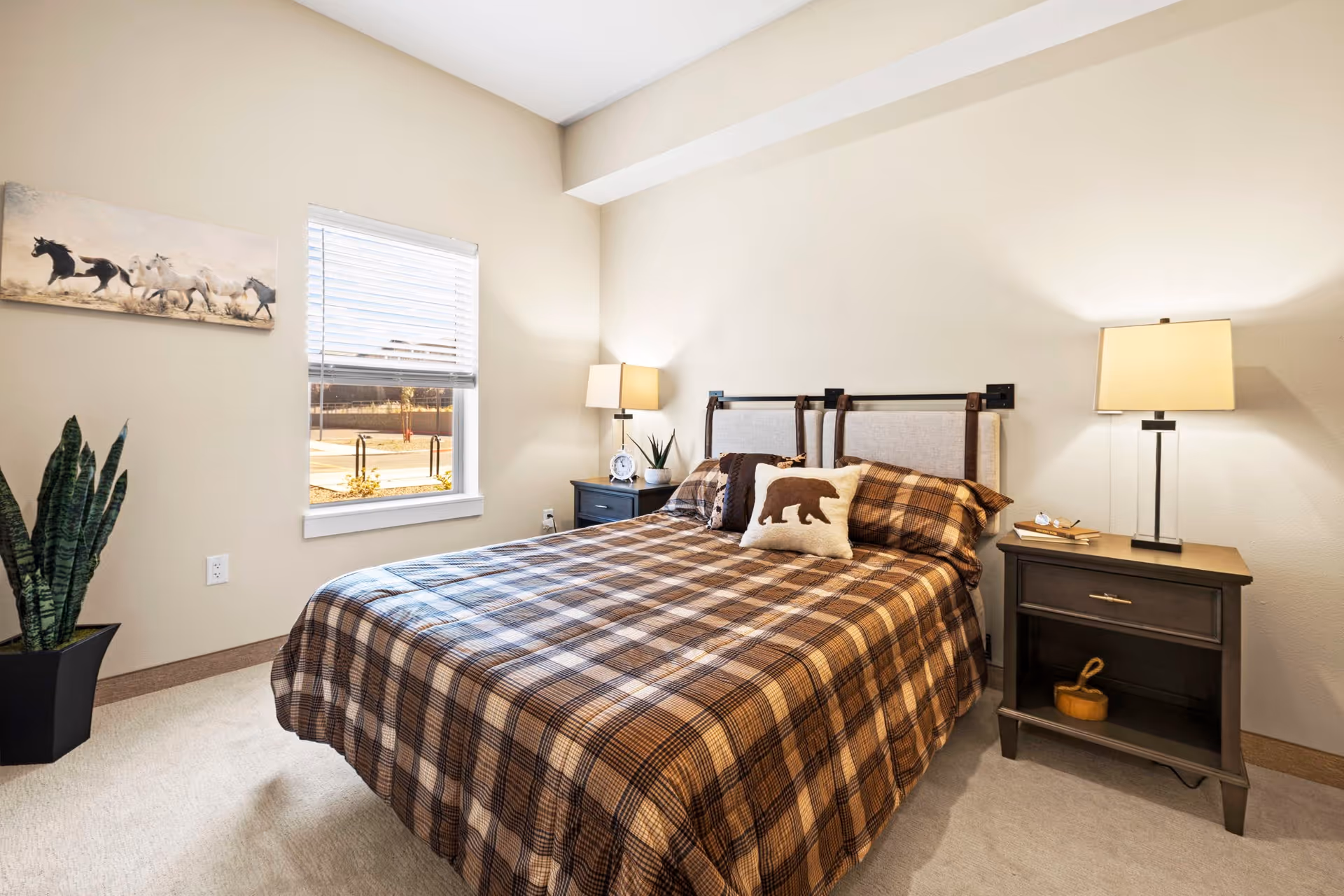 A cozy bedroom with a double bed covered in a brown plaid bedspread and pillows, including one with a bear design. There are two dark wooden nightstands on either side of the bed, each with a lamp. A window with blinds lets in natural light, and a potted plant is placed near the window. A painting of running horses hangs on the wall.