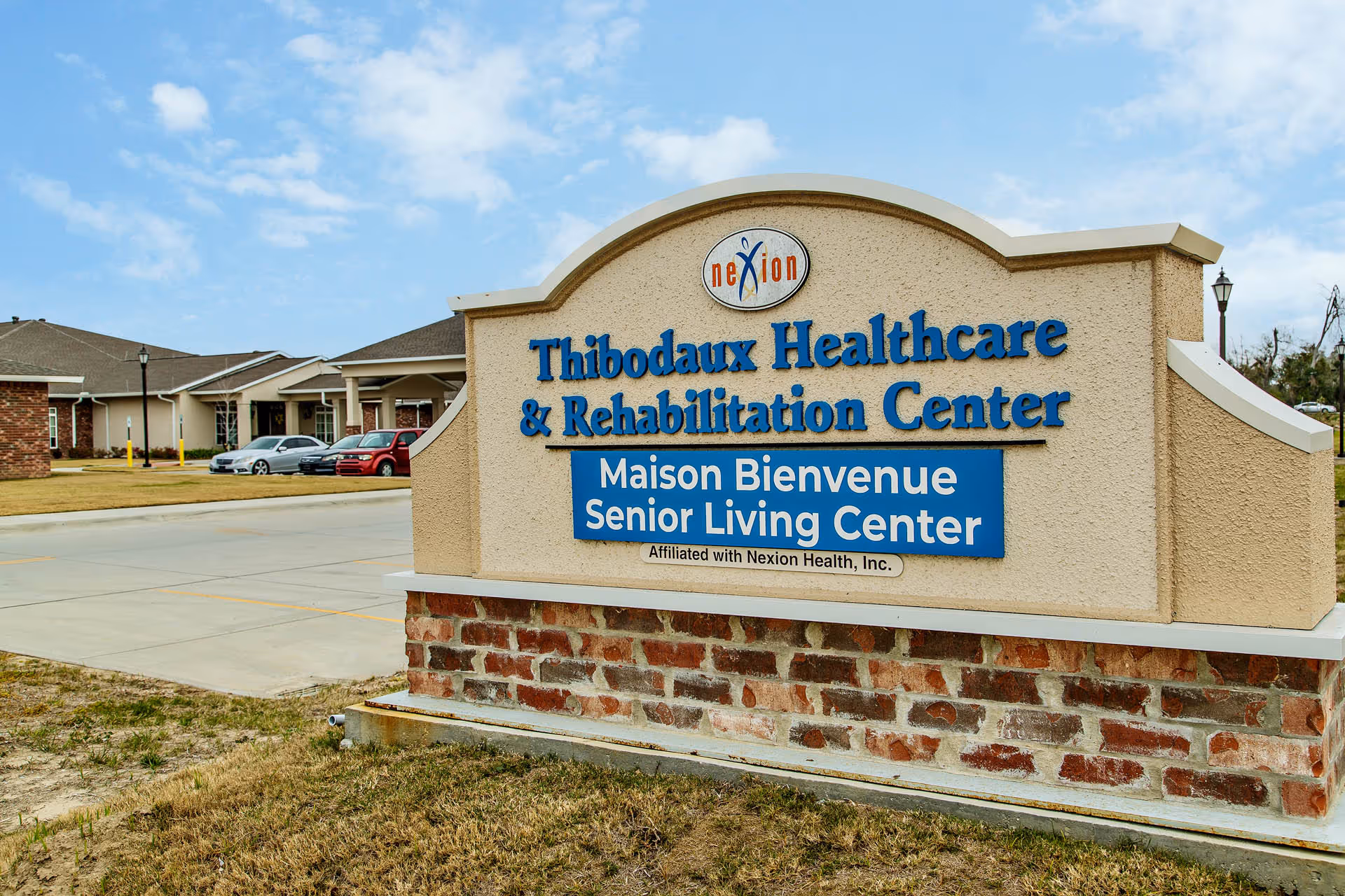 Outdoor view of a sign for Thibodaux Healthcare & Rehabilitation Center and Maison Bienvenue Senior Living Center, with a building and parked cars in the background under a partly cloudy sky.