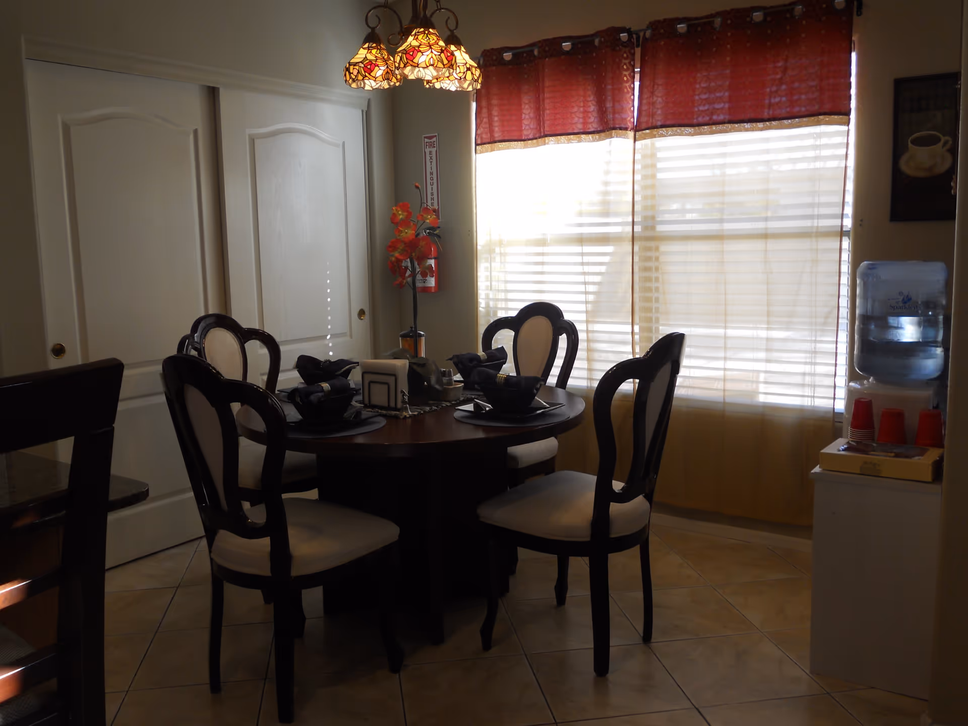 A dining area with a round wooden table set for four with dark napkins and placemats. Four chairs with cream-colored cushions surround the table. A decorative hanging lamp with stained glass shades is above the table. A window with red and beige curtains lets in natural light. A water dispenser with red cups is on a small white cabinet to the right. A fire extinguisher and a tall vase with red flowers are against the wall near two white sliding doors.