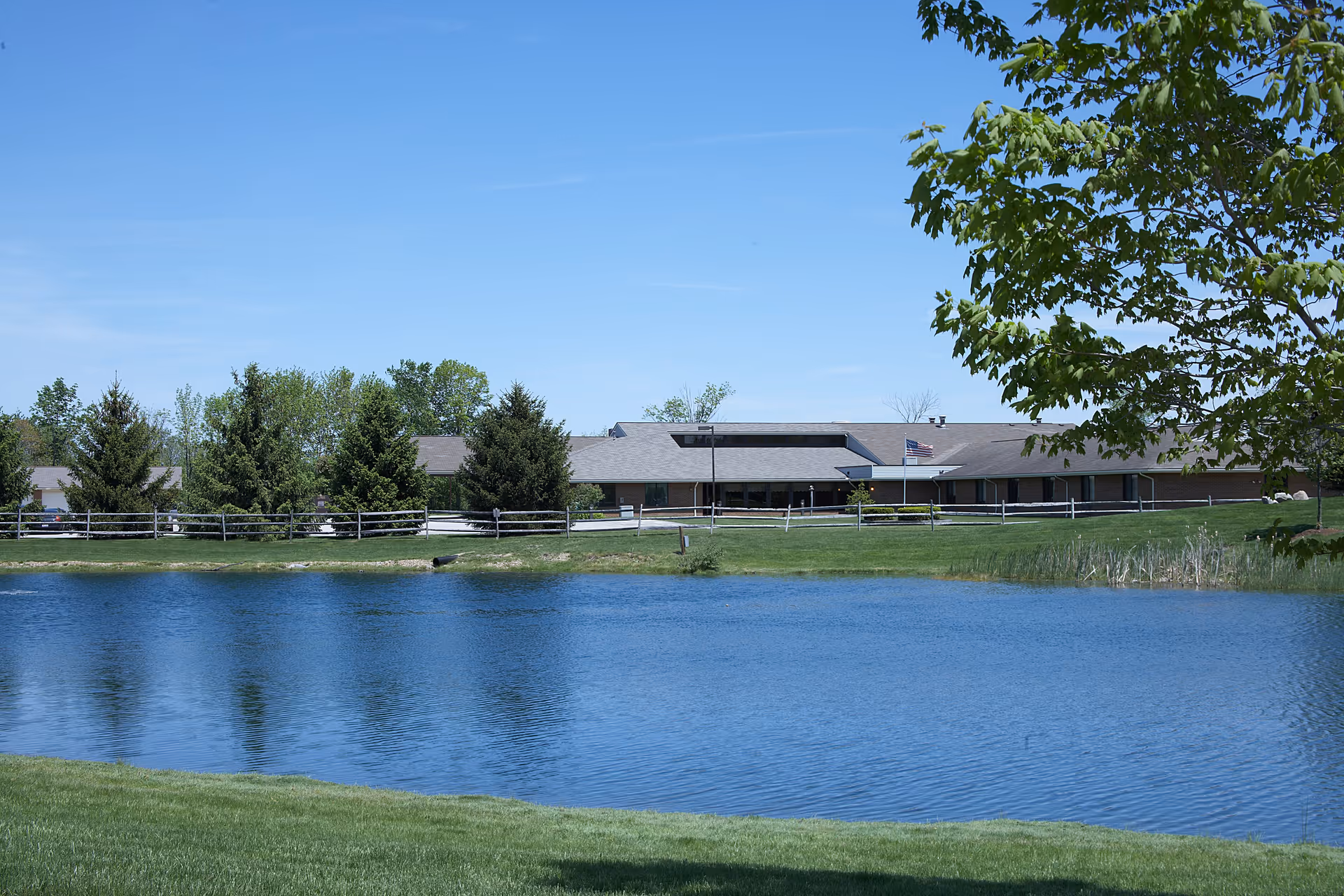 A single-story building with a gray roof and brick walls situated behind a small pond. The scene includes green grass, several trees, a wooden fence, and a clear blue sky. An American flag is visible near the building.