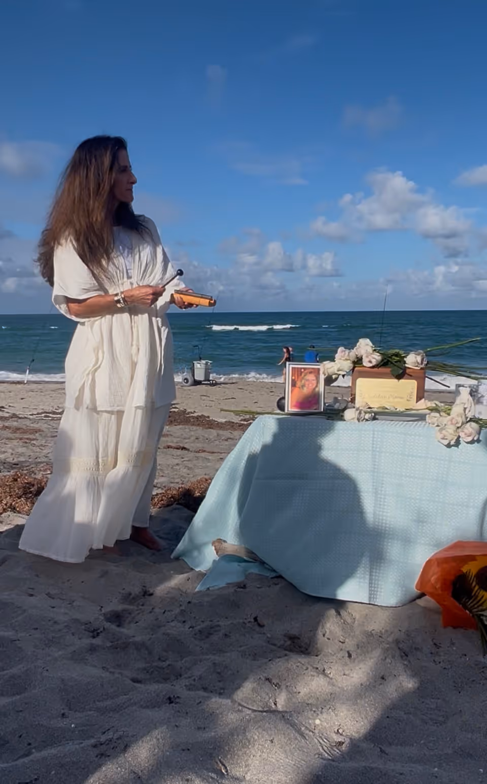 Woman in a white dress stands by a beach memorial table with photos and flowers.