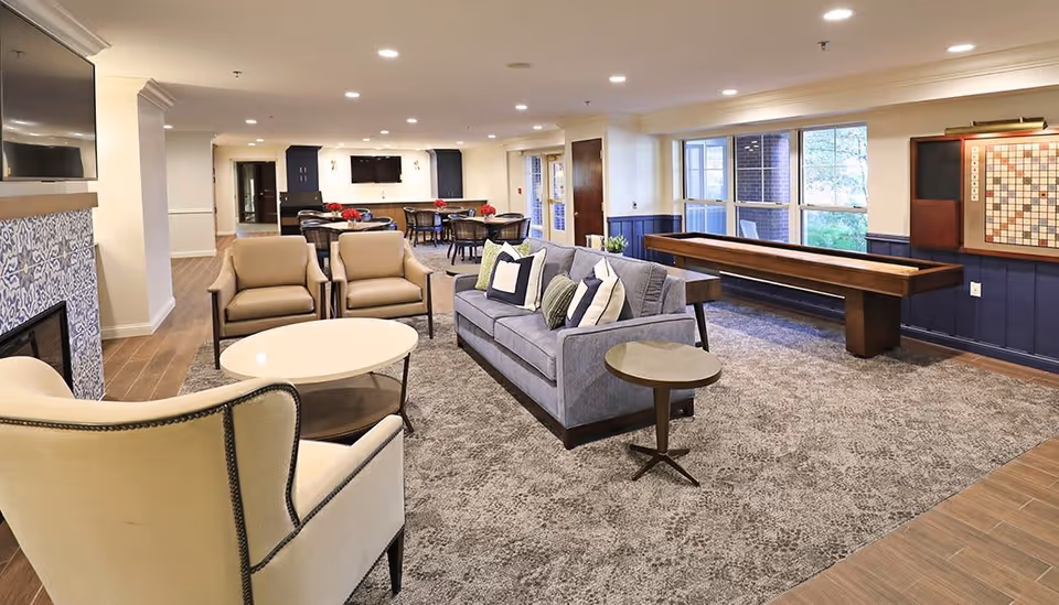 A spacious, well-lit common area with a gray sofa adorned with pillows, two beige armchairs, a round white coffee table, and a small side table. In the background, there are several round tables with chairs, a shuffleboard table near large windows, and a wall-mounted Scrabble board. The room features a fireplace with decorative tile and a mounted TV above it.