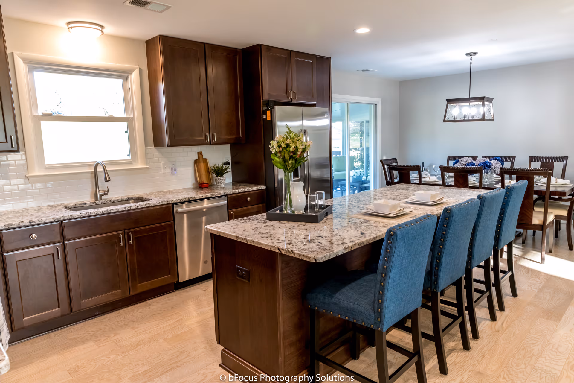 A modern kitchen and dining area featuring dark wood cabinets, a granite countertop island with blue upholstered bar stools, stainless steel appliances, and a dining table set with chairs and a floral centerpiece under a rectangular chandelier.