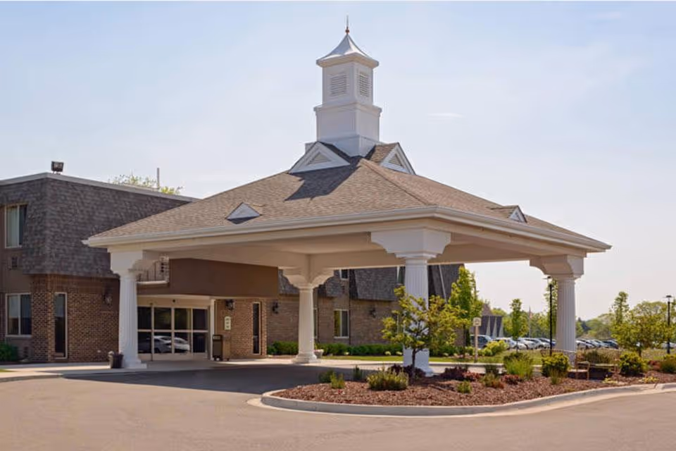 Entrance of a senior living facility with a covered drop-off area supported by white columns, a brick building in the background, landscaped greenery, and a clear sky.