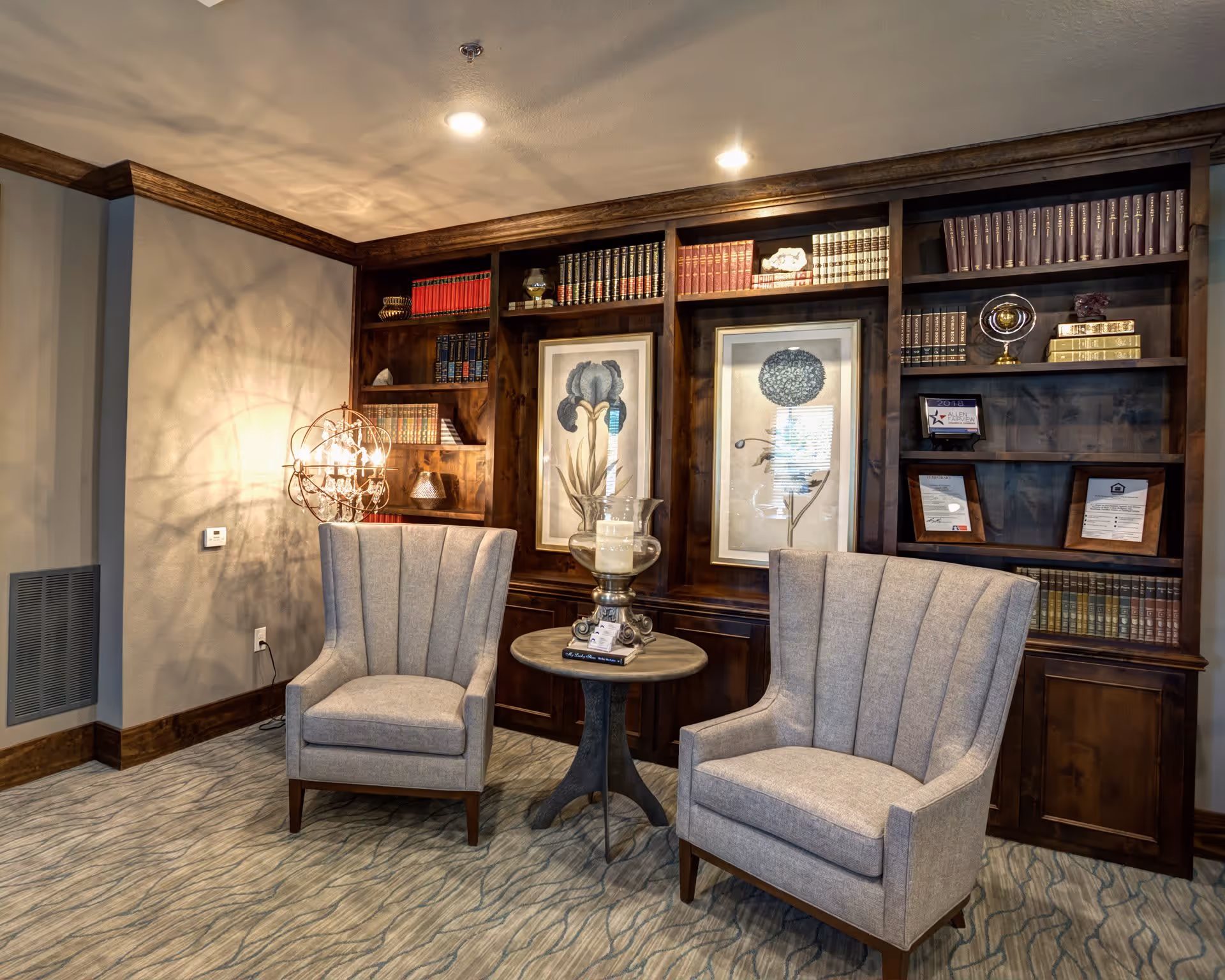 A cozy sitting area with two gray upholstered armchairs facing a small round wooden table. Behind the chairs is a large wooden bookshelf filled with books, framed botanical prints, and decorative items. A unique spherical lamp casts shadows on the wall, and the floor is covered with a patterned carpet.