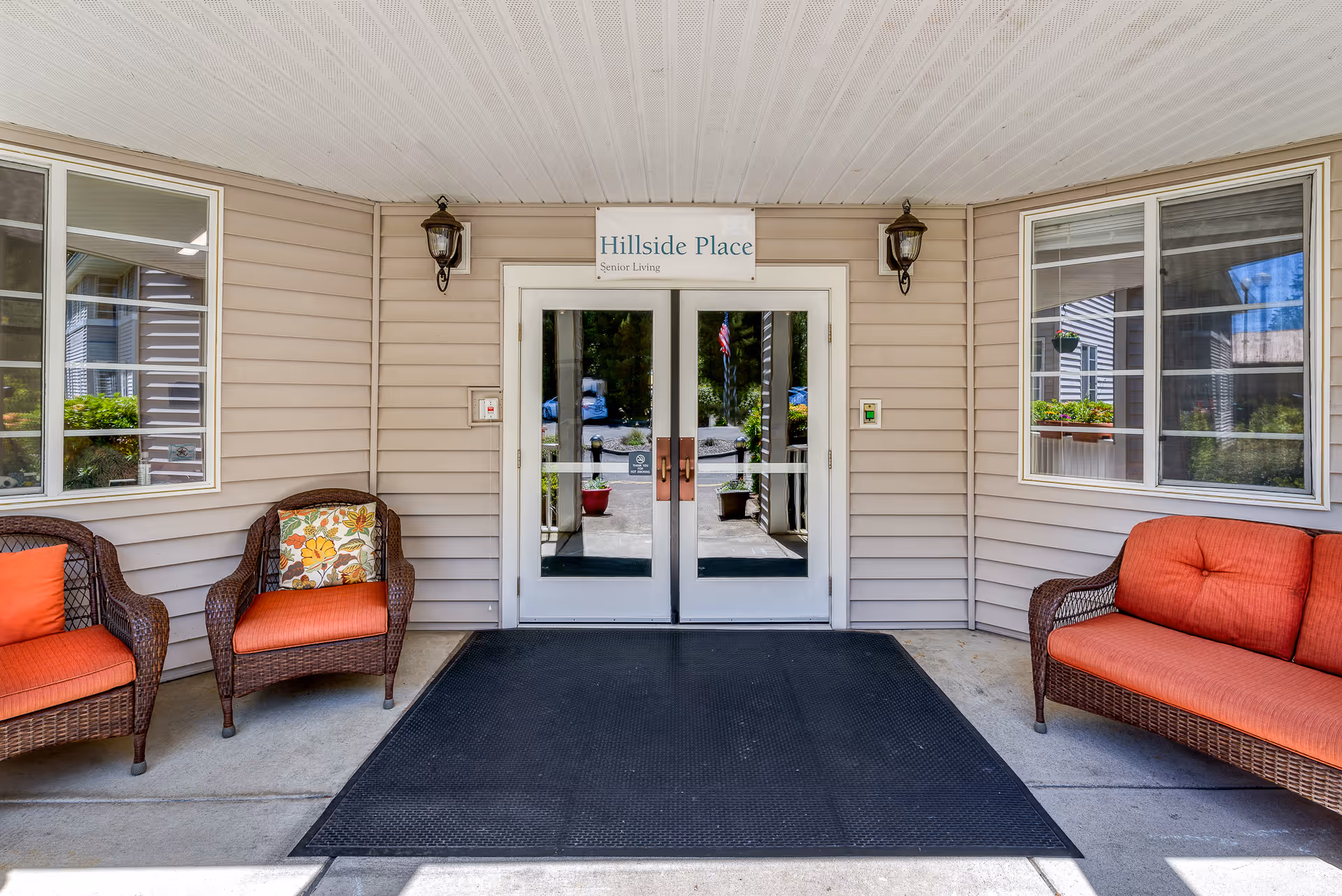 Covered entrance to Hillside Place Senior Living with double glass doors, wicker seating with orange cushions, and a black welcome mat.