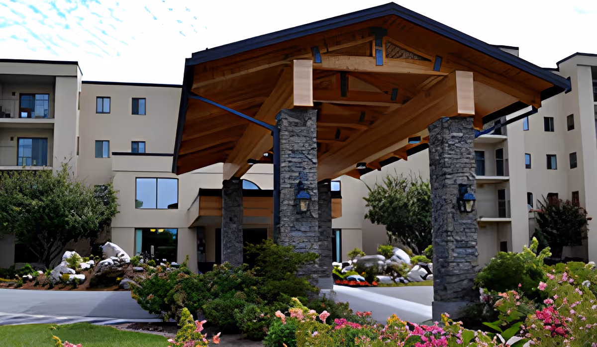 Entrance of Grace Ridge Retirement Community featuring a covered driveway with stone pillars and wooden roof beams, surrounded by landscaped greenery and flowers, with the multi-story building in the background.