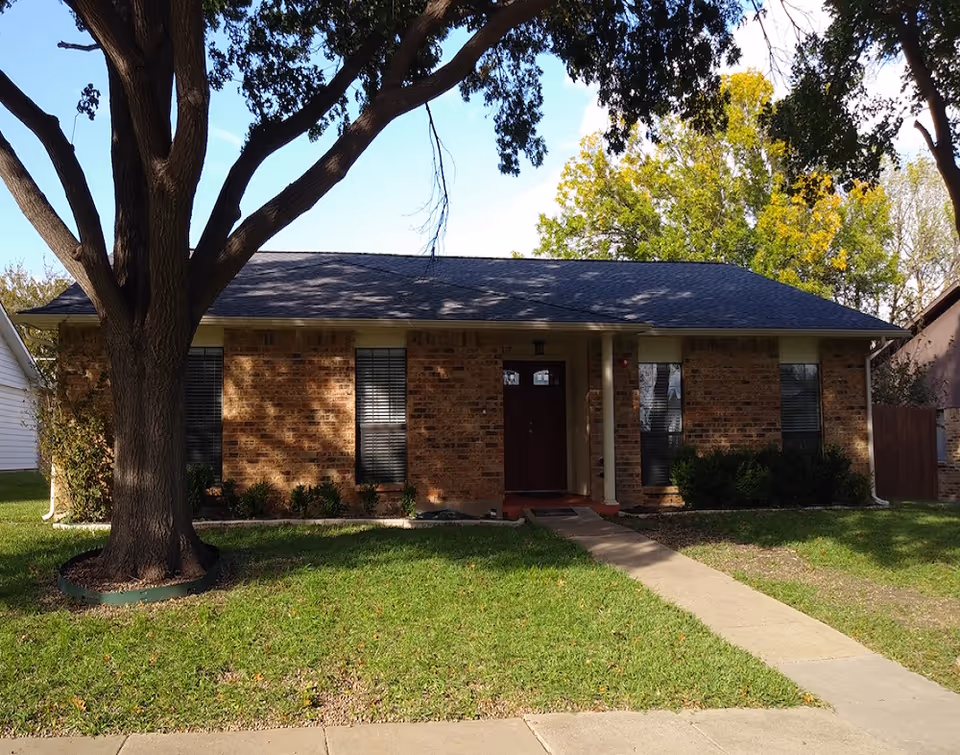 Brick, single-story residential building with a front path, lawn, and a large tree shading the entry.
