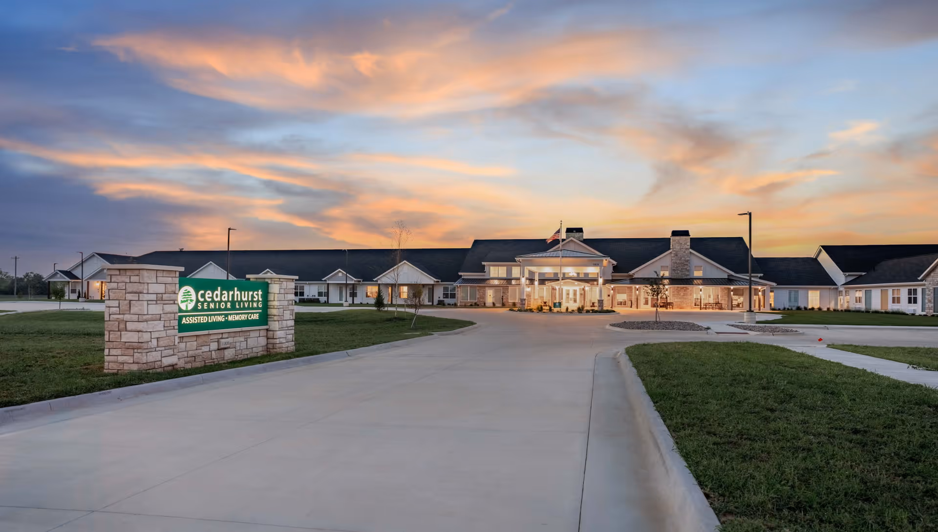 Exterior view of Cedarhurst Senior Living of Wichita at sunset, showing a large building with a covered entrance, well-lit windows, and a stone sign in the foreground displaying the facility's name and services including assisted living and memory care.